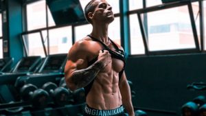 Muscular man showing abs and tattoos in a gym setting.